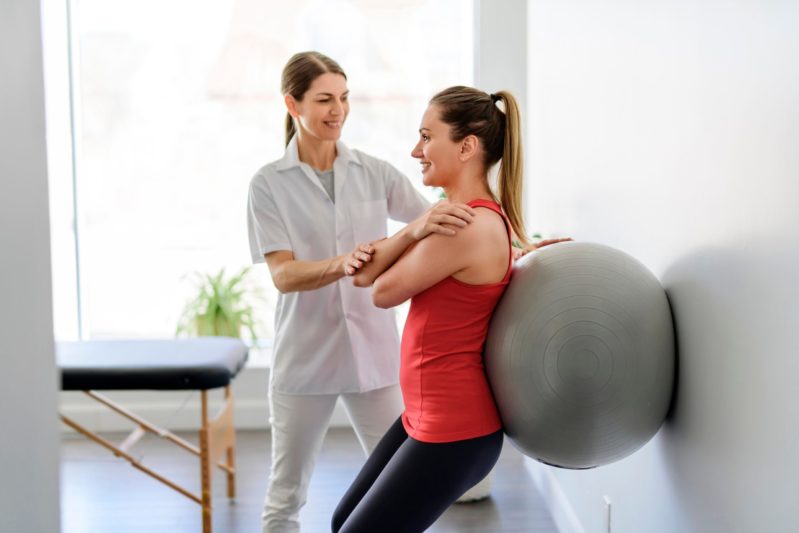 Fisioterapeuta ayudando a una mujer a hacer estiramiento en pared con balón de pilates